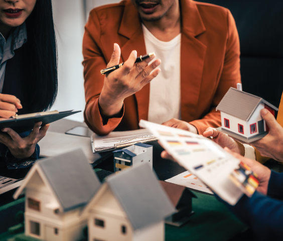 Pessoas discutindo numa mesa de trabalho com vários papéis em volta e com um modelo de casa em miniatura